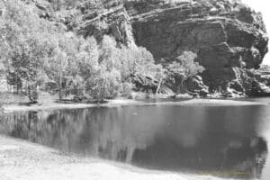 Black and white photograph of a large watering hole between two large rocky outcrops.