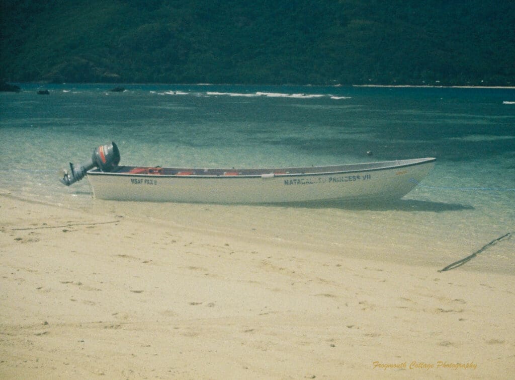 Photograph of a long speed boat with faded writing along the hull. The boat is sitting at the edge of the beach with blue ocean behind it and lush green mountains in the background.