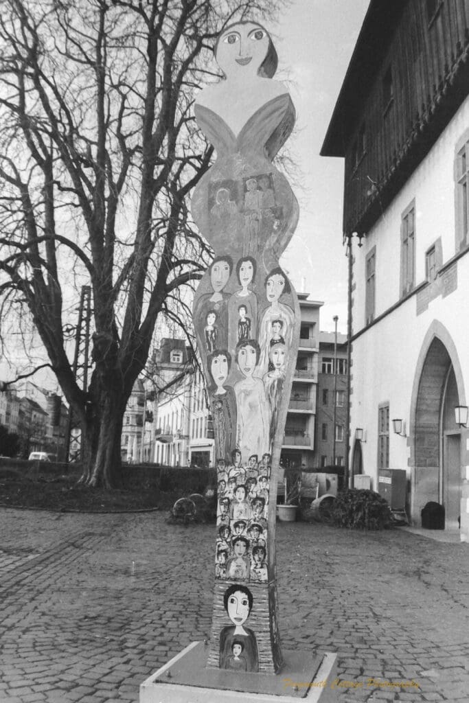 Black and white photograph of a sculpture in the shape of a woman with faces of women painted on it.