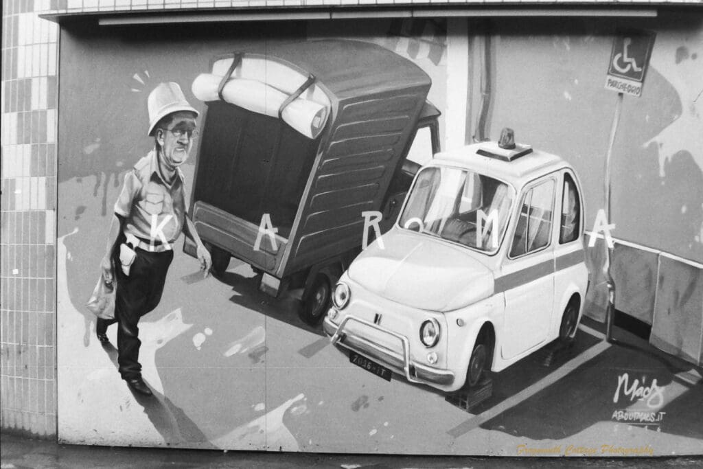 Black and white photograph of a mural painted on a wall. The mural shows a car parked in a disabled parking spot with it's tyres clamped and the word "Karma".