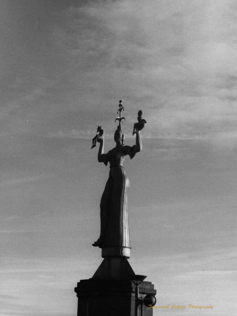 Black and white photograph of a concrete statue of a woman wearing a fools cap on her head and holding her arms up. On her hands sit two small figures.