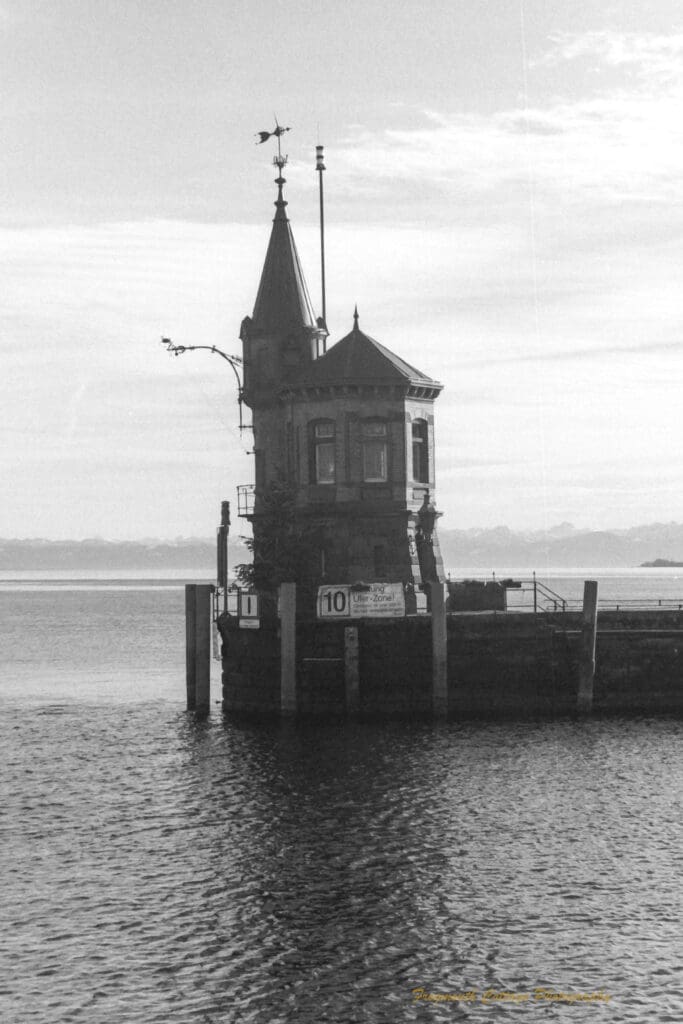 Black and white photograph of a large lake with a jetty in the middle-ground. At the end of the jetty is a small building with a weather vane on top.