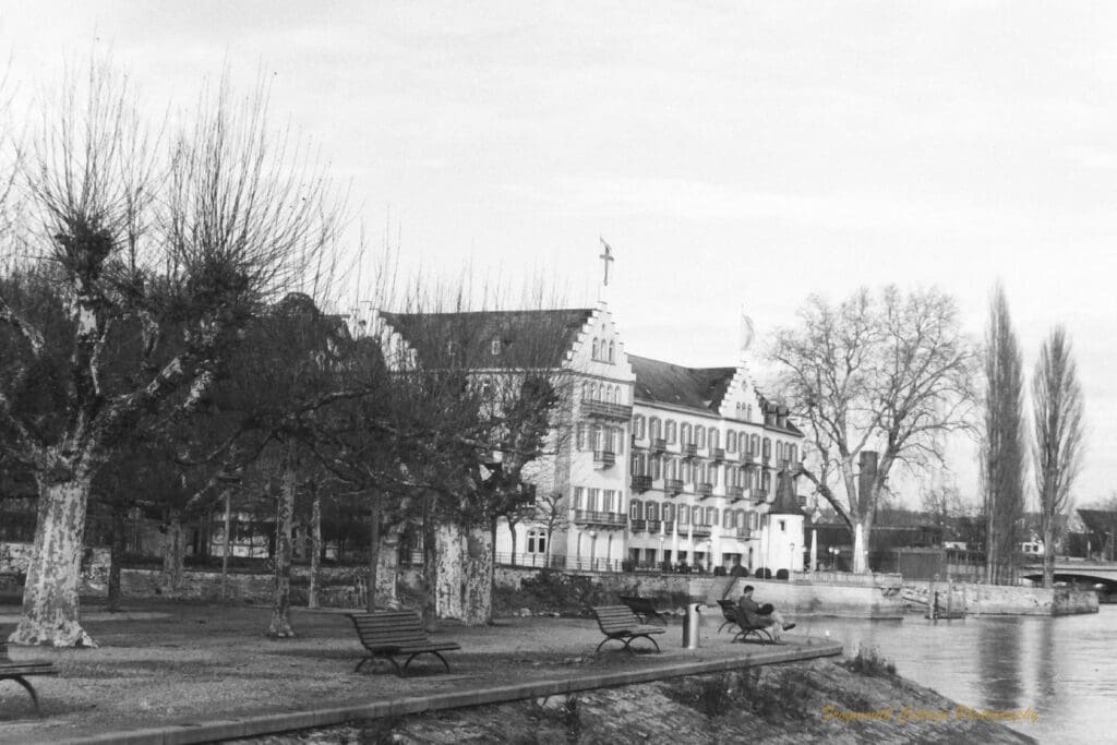 Black and white photograph of a park and buildings along the edge of lake, a man is sitting on park bench