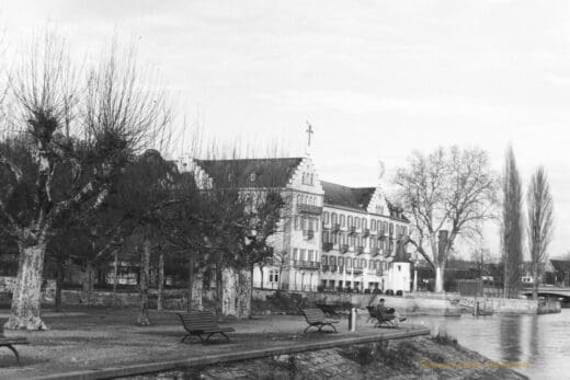 Black and white photograph of a park and buildings along the edge of lake, a man is sitting on park bench