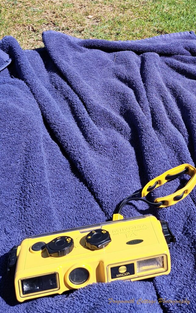 Photograph of the Minolta Weathermatic A lying on a blue towel placed on grass, with a pool in the background.