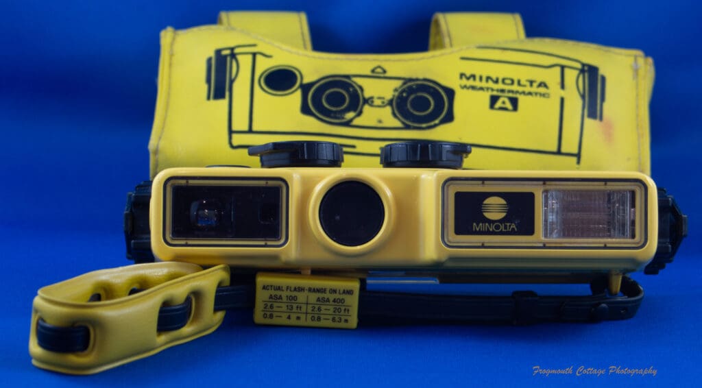 Photograph of the Minolta Weathermatic A withwrist strap and the wrap-around case behind it.