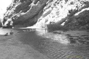 Black and white photo of a boy walking across stepping stones across a stream of water