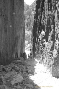 Black and white photo of two people standing between two sheer walls of rock with another rocky outcrop in the background. The sun is shining through the gap between the walls.