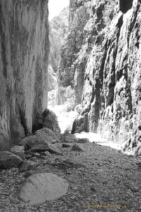 Black and white photo looking along a chasm between two cliff faces, the ground is rocky and there is a shaft of light shining through the far end. There are more rocky outcrops in the distance.