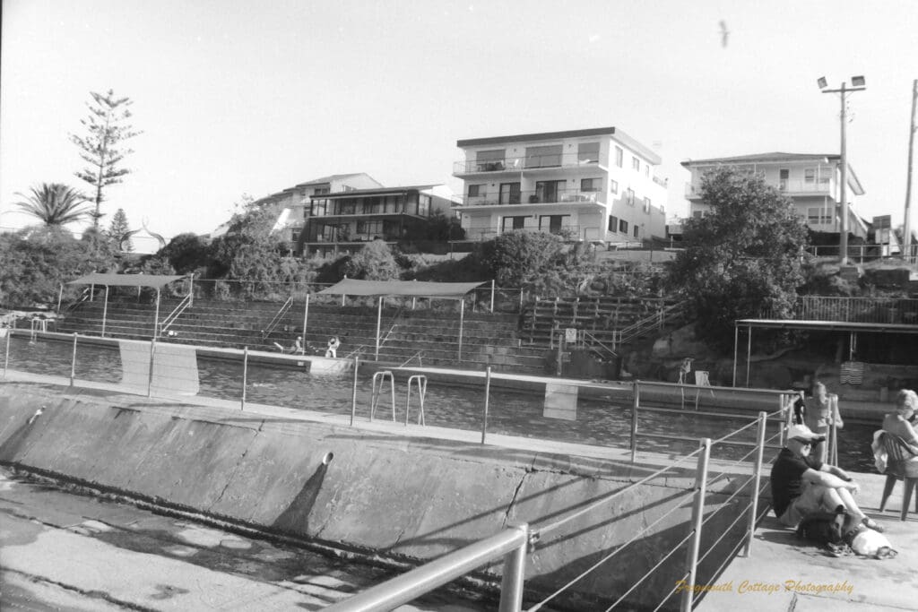 Black and white photograph of a swimming pool in front of a row of house with concrete steps leading down to the pool. On the right hand side of the pool people are sitting on the ground an in chairs.