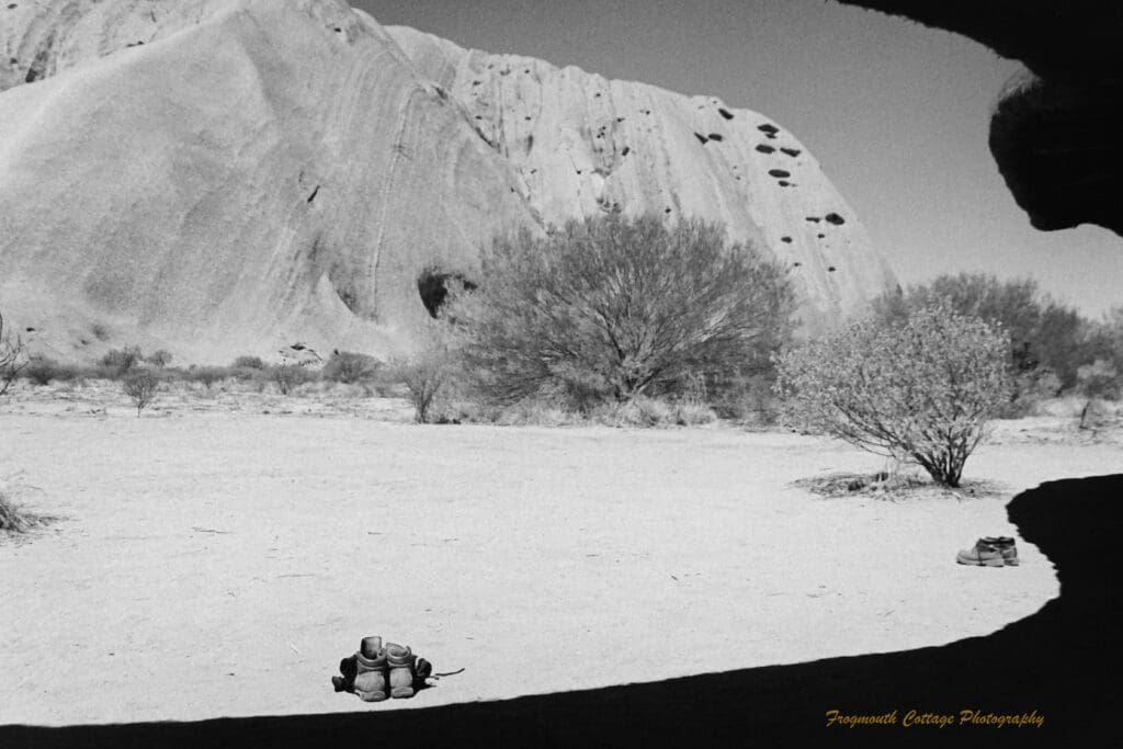 Black and white photo taken in the shadow of a sheltered area. The roof has created a curved shadow on the ground. Two pairs of boots are sitting on the ground in front. The side of Uluru is in the background.