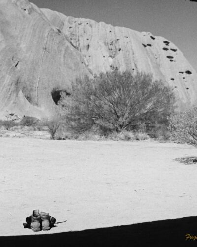 Black and white photo taken in the shadow of a sheltered area. The roof has created a curved shadow on the ground. Two pairs of boots are sitting on the ground in front. The side of Uluru is in the background.
