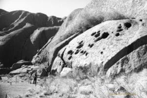 Black and white photo of Uluru in the Uluru and Kata Tjuta National Park, Northern Territory. There are two people standing at the base of the rock, they are very small in comparision to Uluru.