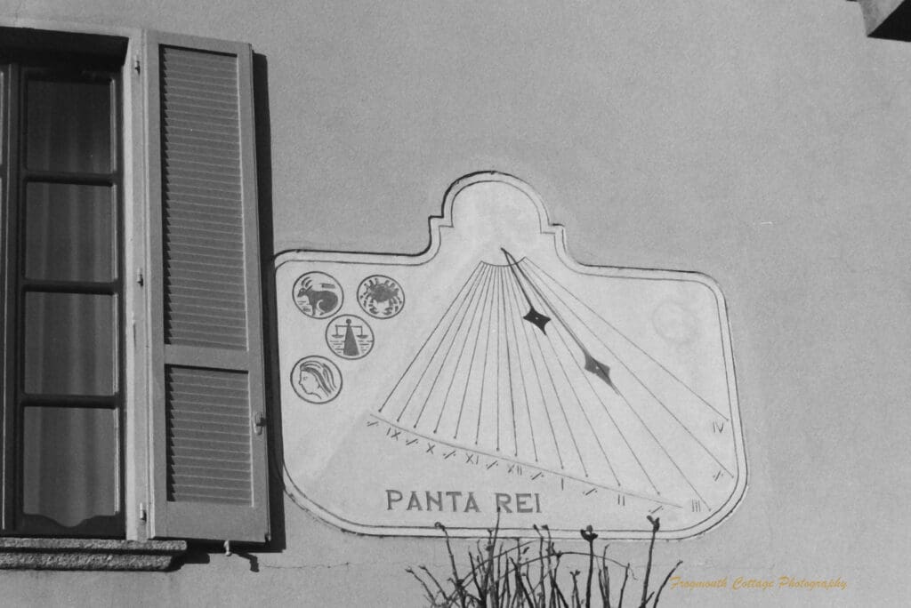 Black and white photo of the exterior stone wall of a house with a window on the left hand side. The focus of the photo is the sun dial on the wall with the words "Panta Rei"on it.