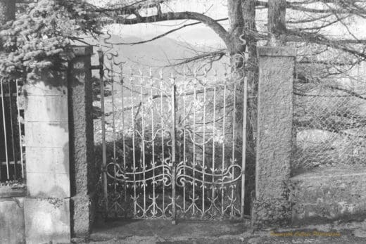 Black and white photograph of a wrought iron gate in a fence with the view of a valley behind it.