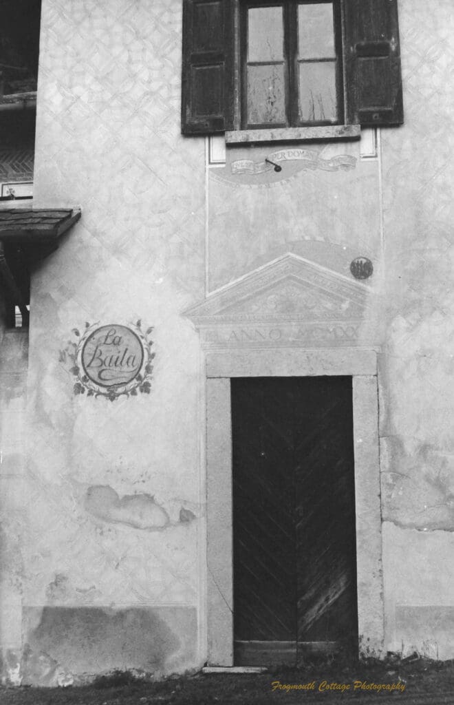 Black and white photograph of the wooden door of a stone building, there is a window above the door with shutters and a an engraving to the side that says "La Baila".