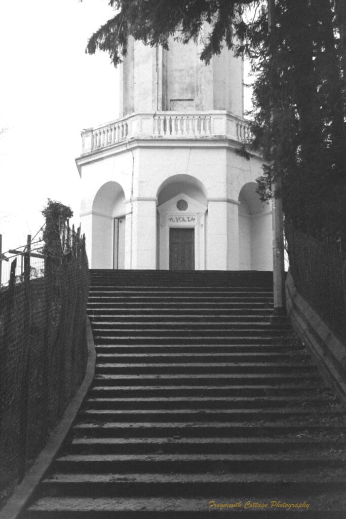 Black and white photograph of stone stairs leading to a white octagonal building, above the door a sign reads "A Volta".