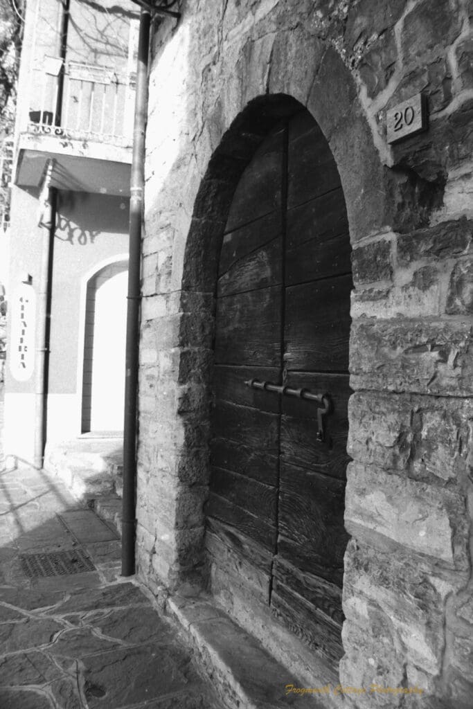 Black and white photograph of a wooden door set into a stone building, there is a large metal slider across the door, and the number 20 to the side.