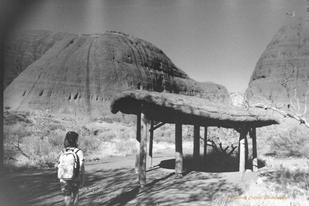 Black and white photo of a a boy walking towards a thatched shelter which sits in front of two very large rocks that tower above him