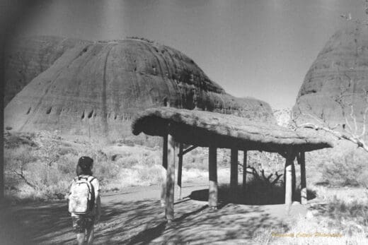 Black and white photo of a a boy walking towards a thatched shelter which sits in front of two very large rocks that tower above him