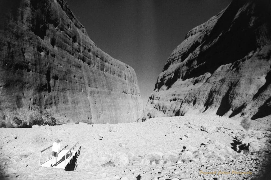 Black and white photograph of two very long large rock formations and a boy walking over a small bridge away from the gap in between